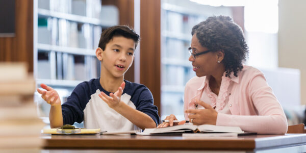 A serious middle school age boy sits at a table with a mature female tutor in his school library.  He gestures as he tells her his issues with the homework.